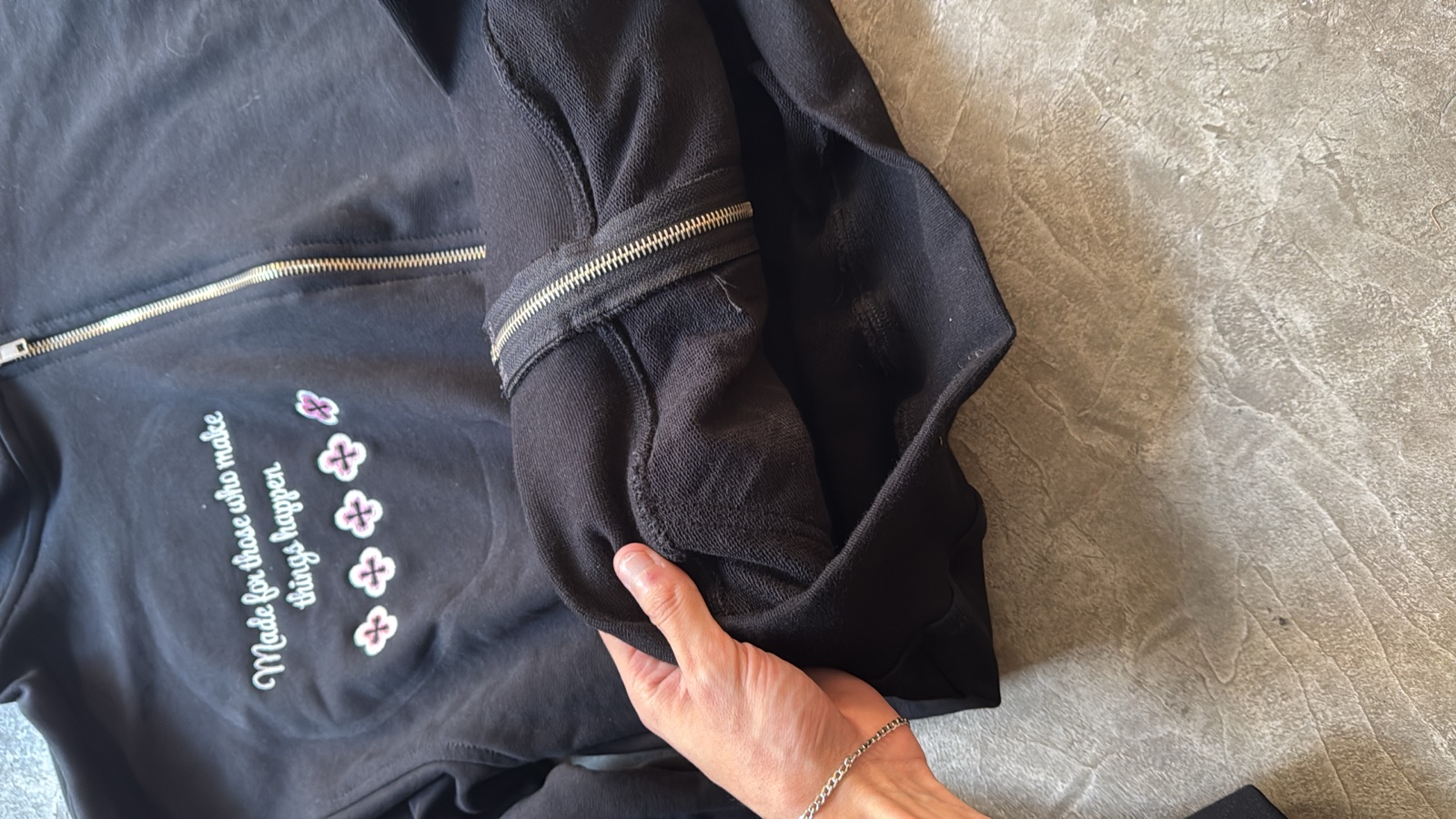 Black zip-up jacket with embroidered text and floral motifs on sleeve hand inspecting metal zipper cuff detail