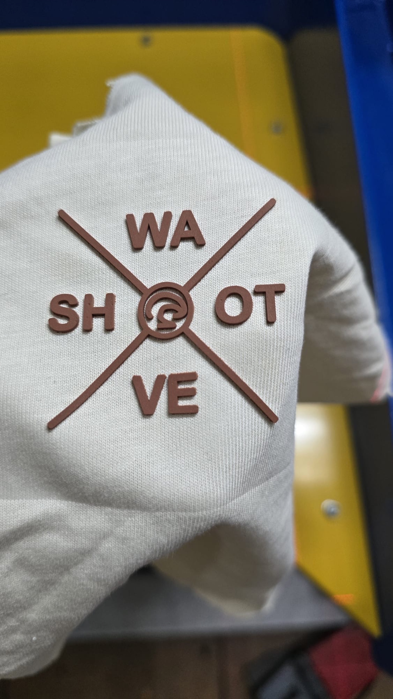 Close-up of off-white garment with raised brick-print Shot Wave compass logo in brown on heat press