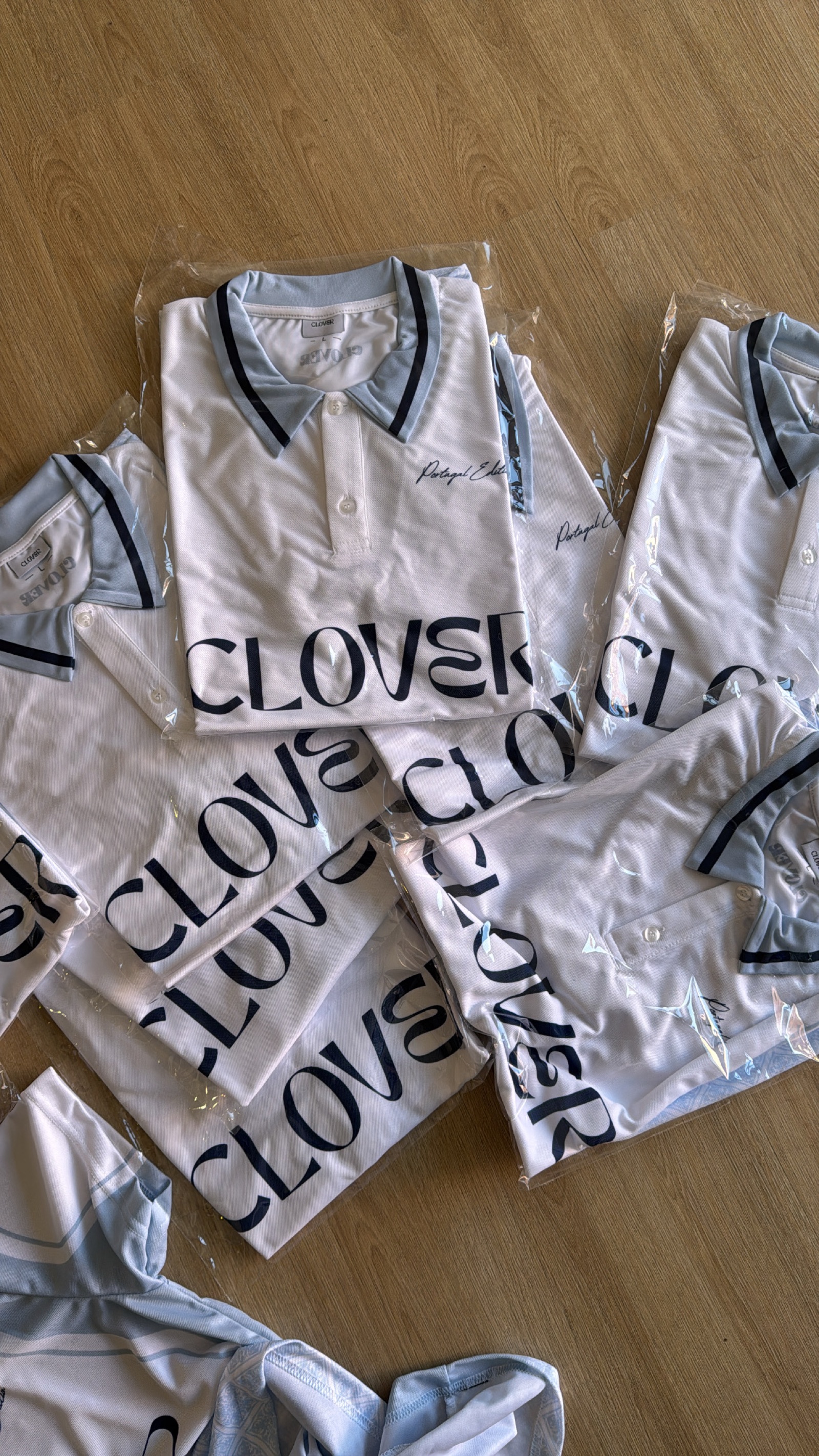 Multiple white Clover branded polo shirts with navy trim and script embroidery laid out on wooden surface