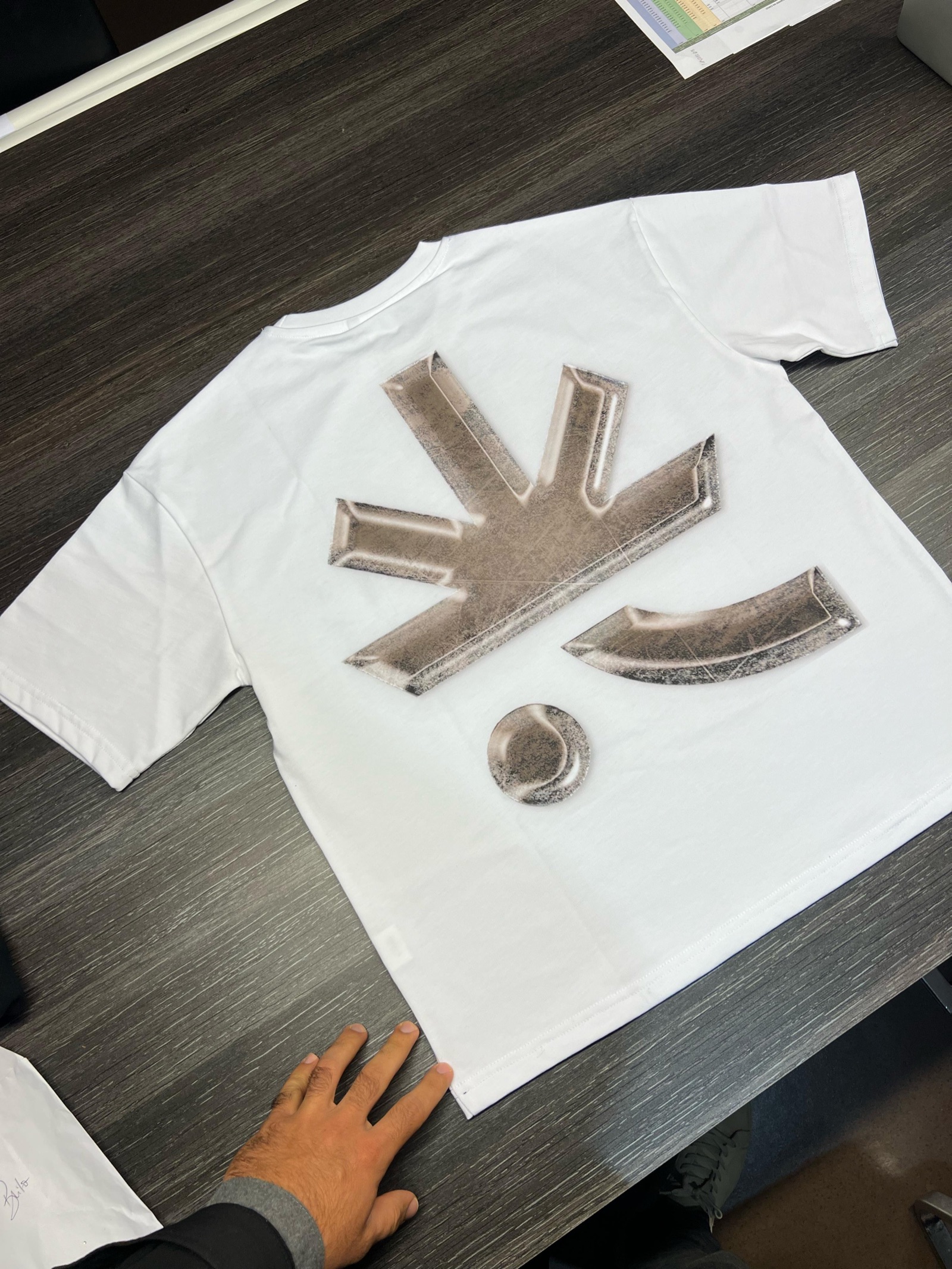 White oversized t-shirt laid on work table showing large metallic silver abstract logo print on back