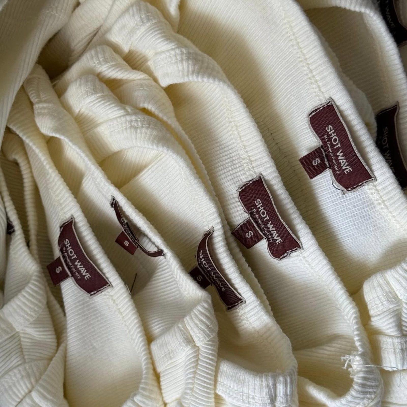 Pile of off-white ribbed tank tops with burgundy Shot Wave branded woven size labels