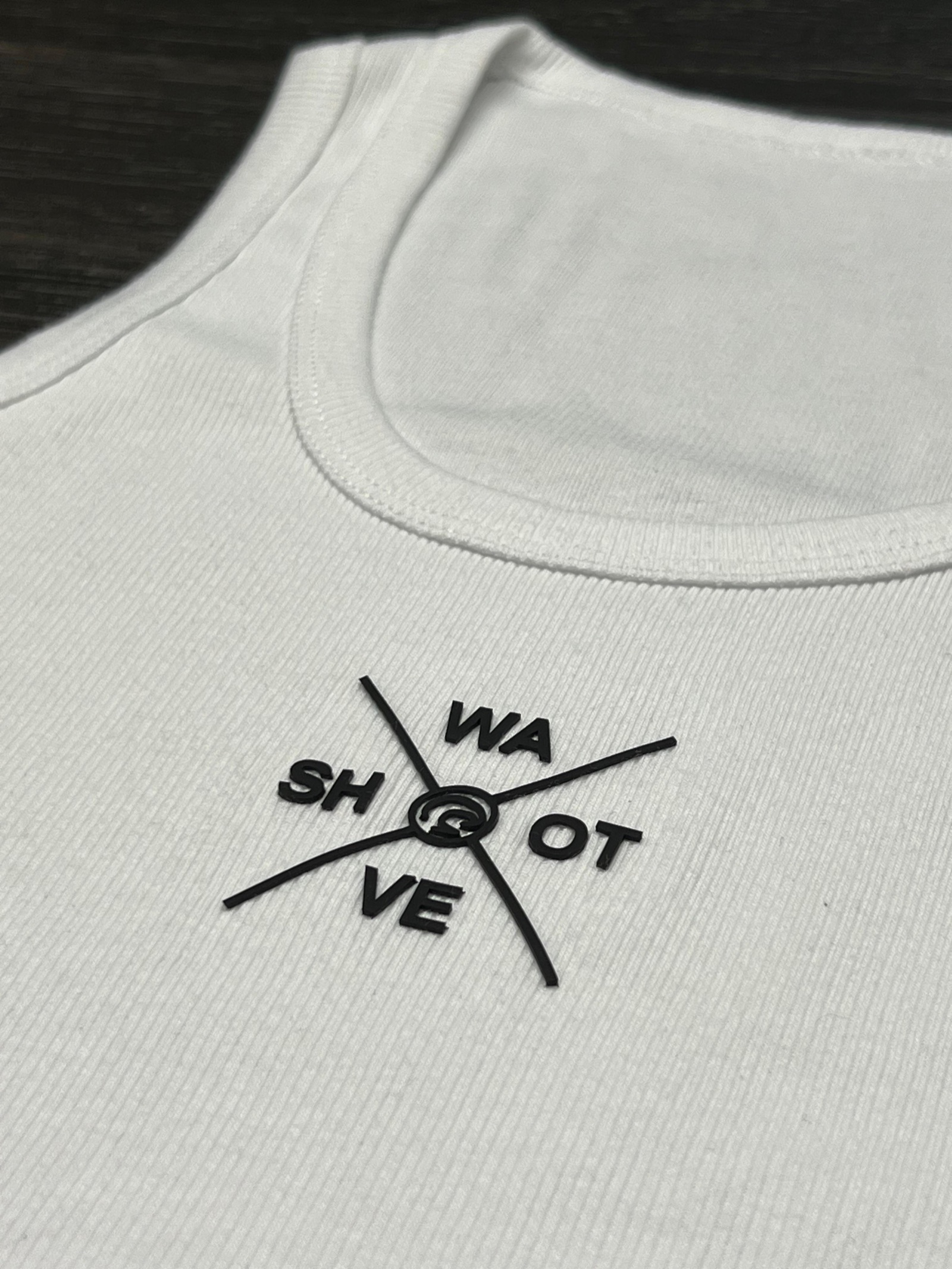 White ribbed tank top with black brick-print Shot Wave compass logo on chest against dark surface
