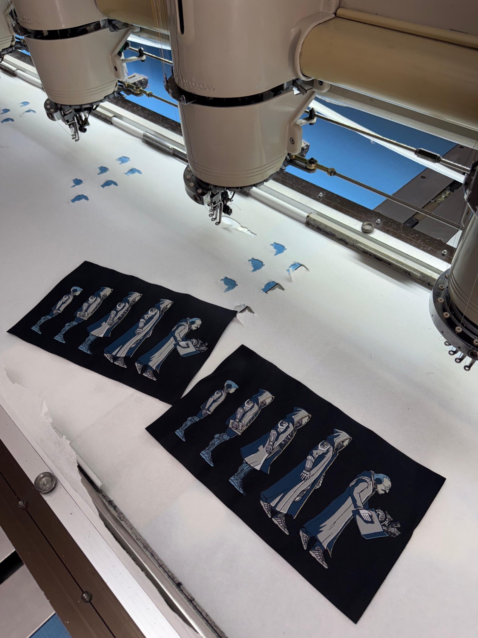 Industrial multi-head embroidery machine stitching blue thread details onto dark fabric patches with figure artwork