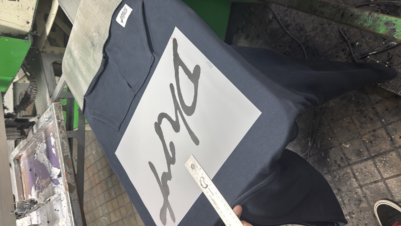 Dark navy t-shirt on screen-printing press platen showing large white cursive script graphic being applied