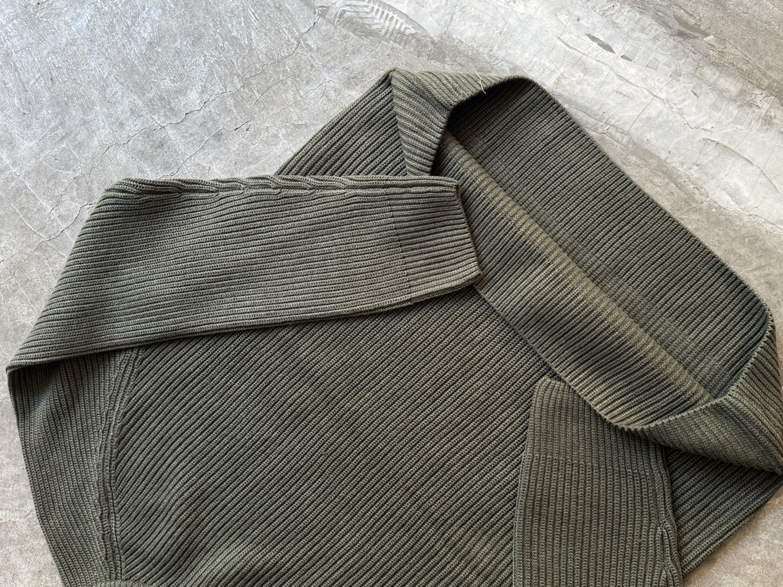 Olive green ribbed-knit sweater laid on concrete surface showing textured ribbing and heavy-gauge knit construction