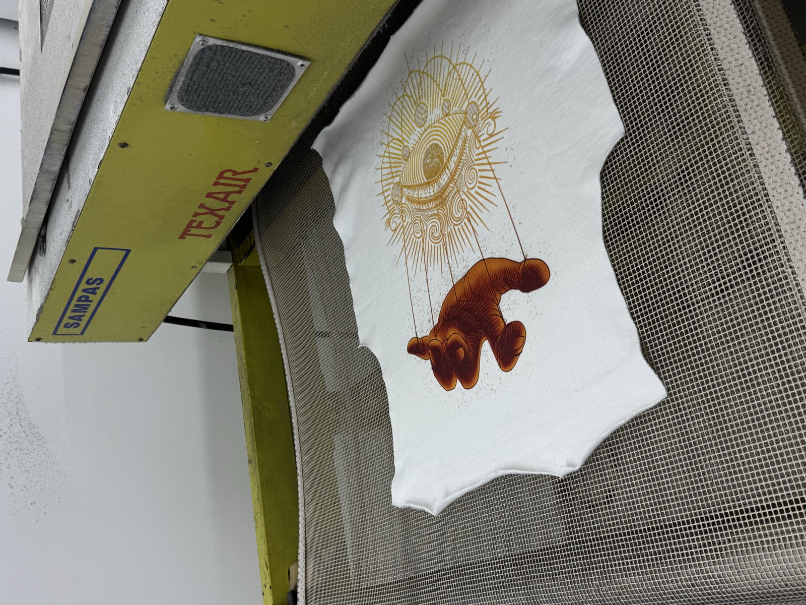 White t-shirt on textile heat press displaying golden-toned illustration of marionette puppet with radiating lines