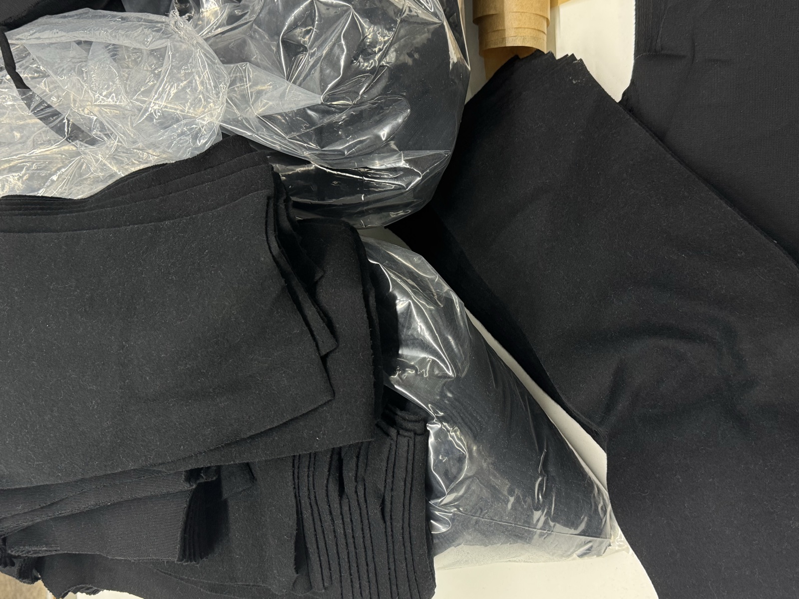 Assortment of black garments including knit ribbed pieces and woven fabrics partially wrapped in plastic on factory surface