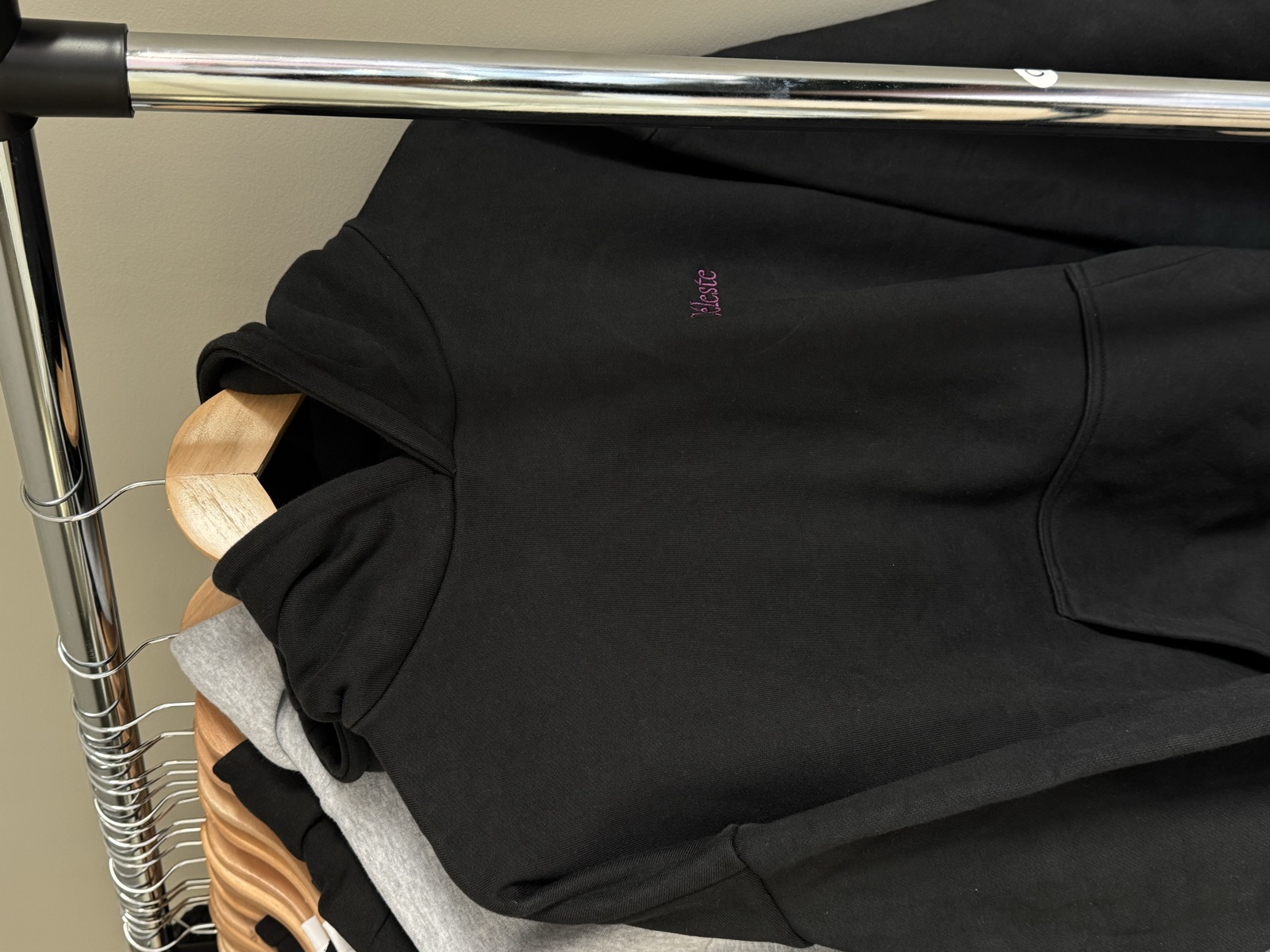 Black hoodie with small embroidered logo on chest hanging on wooden hanger on metal clothing rack