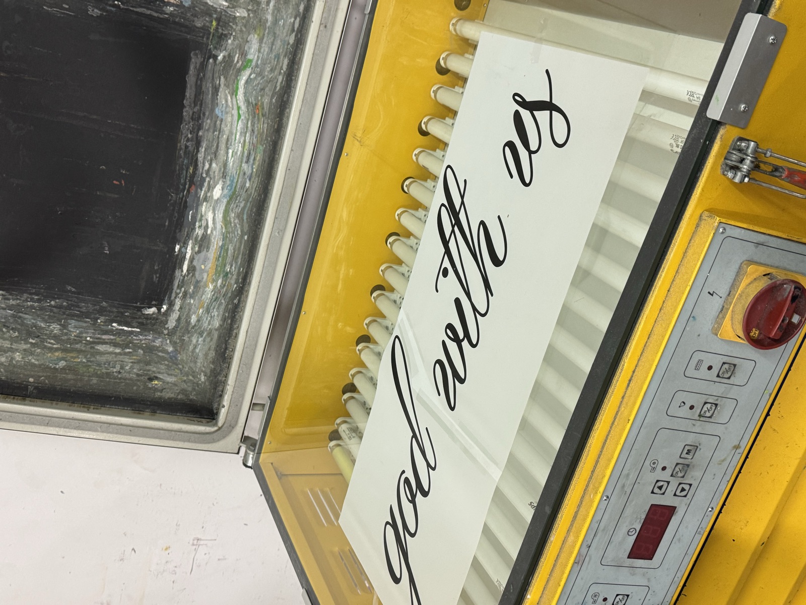 Screen printing frame with god with us cursive text loaded on yellow heat press machine in factory