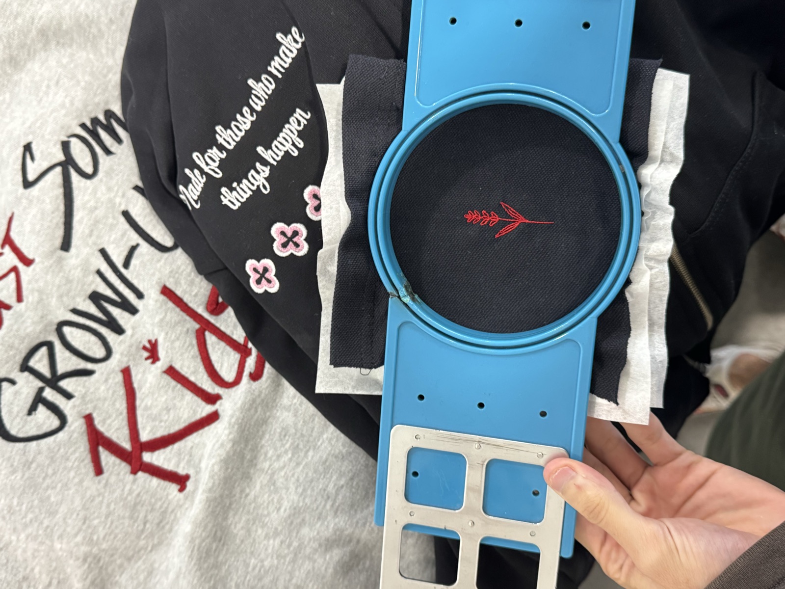 Close-up of embroidery hoop on machine stitching small red arrow logo onto black fabric with printed garments beneath