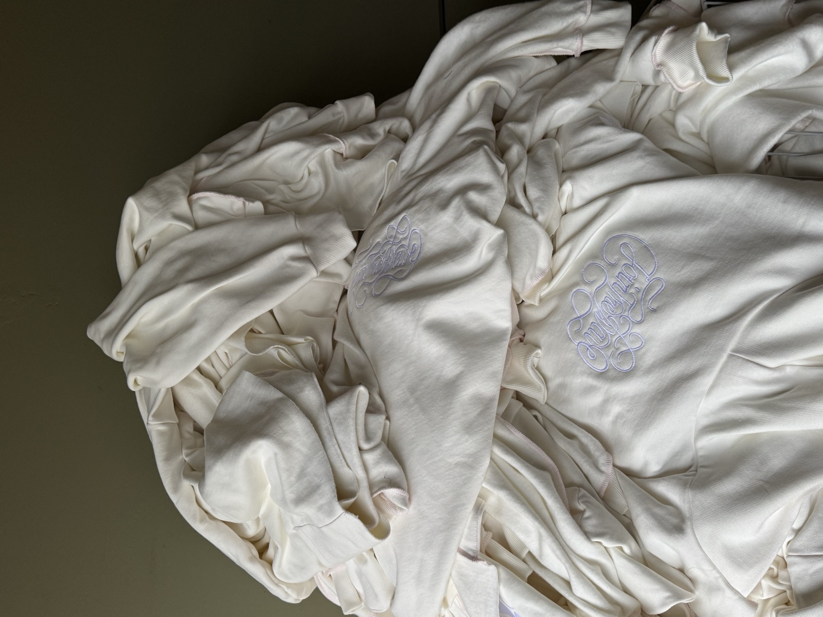 Pile of off-white cotton hoodies with tonal embroidered crest logo visible on the chest area