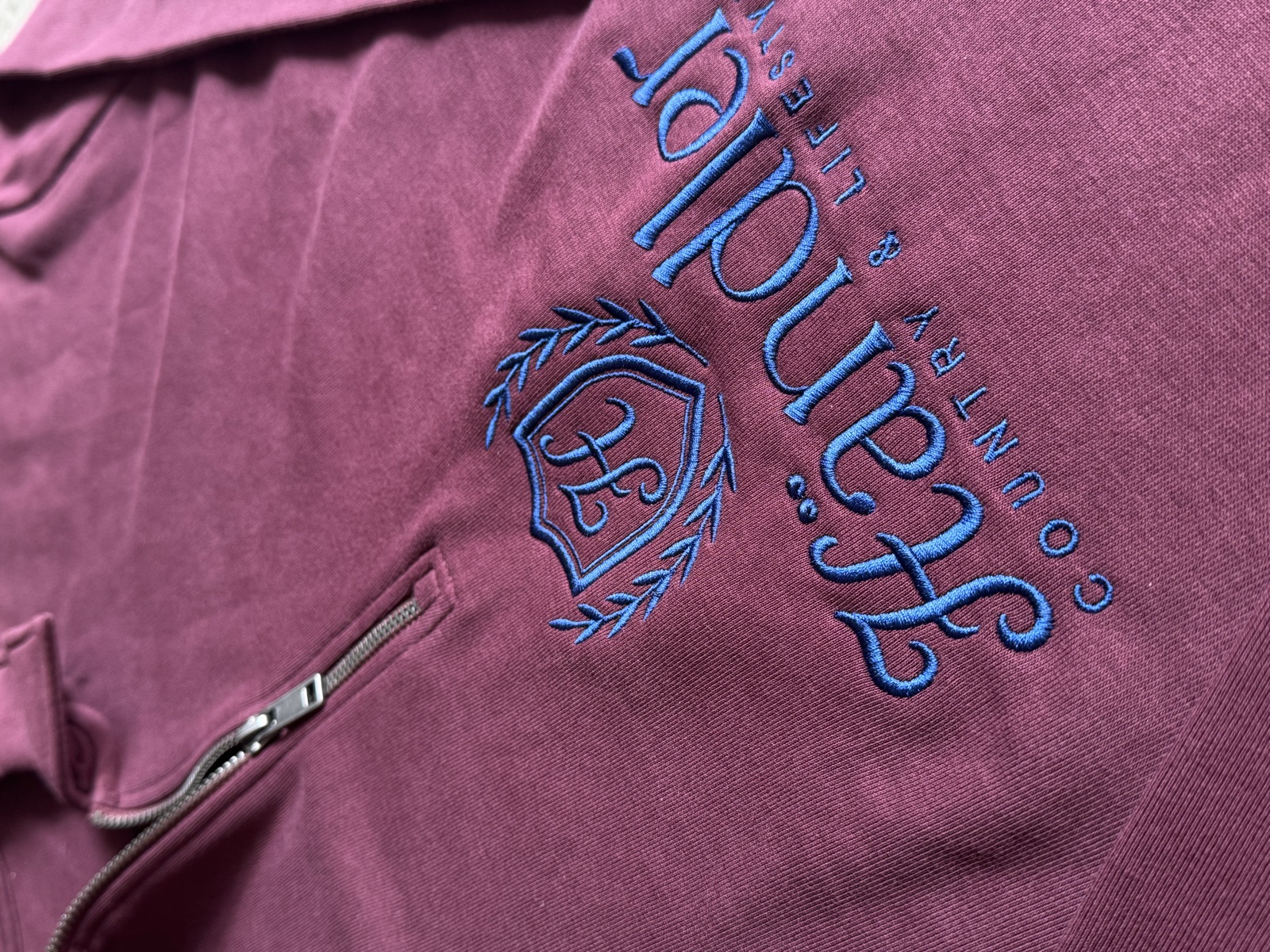 Close-up of burgundy hoodie with blue embroidered Scandale Country & Lifestyle branding and quarter-zip