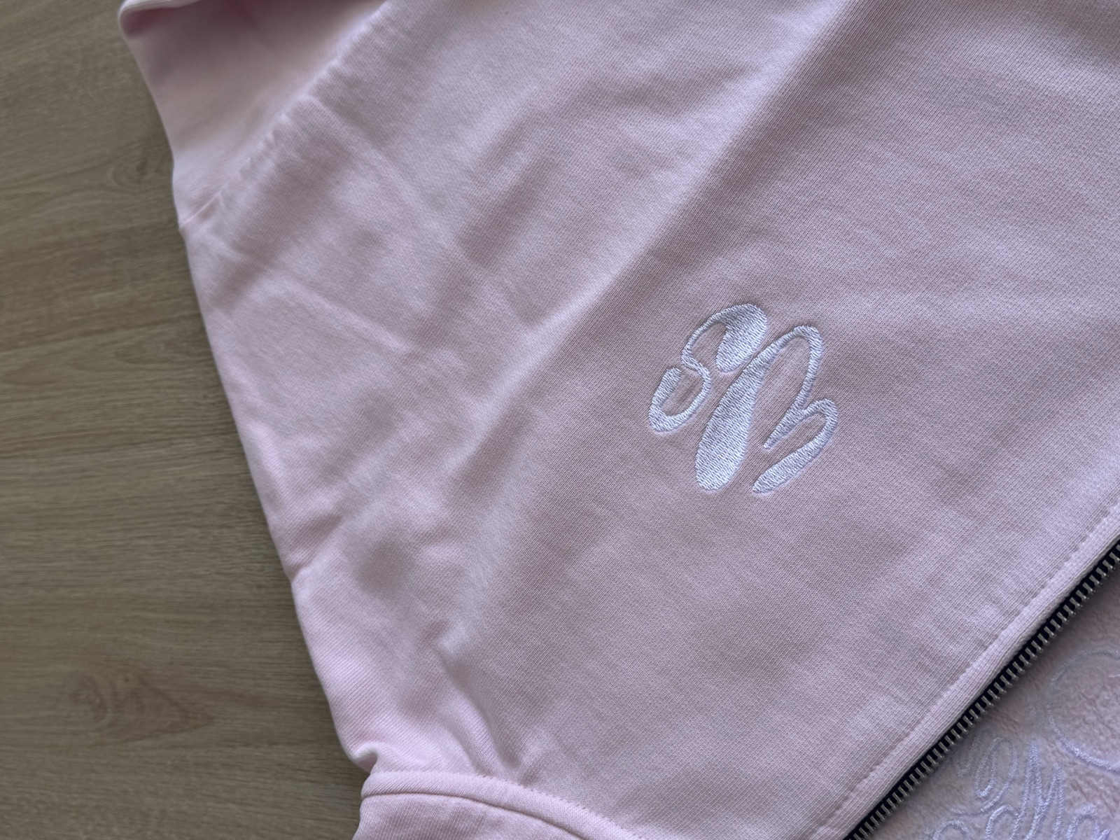 Light pink zip-up hoodie with tonal white embroidered monogram logo on chest laid flat on wooden surface