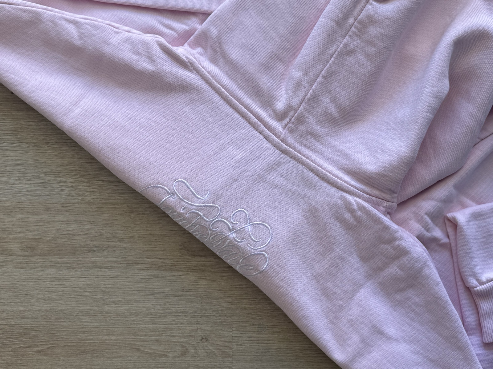 Light pink hoodie sleeve detail showing tonal embroidered script logo on forearm with kangaroo pocket visible