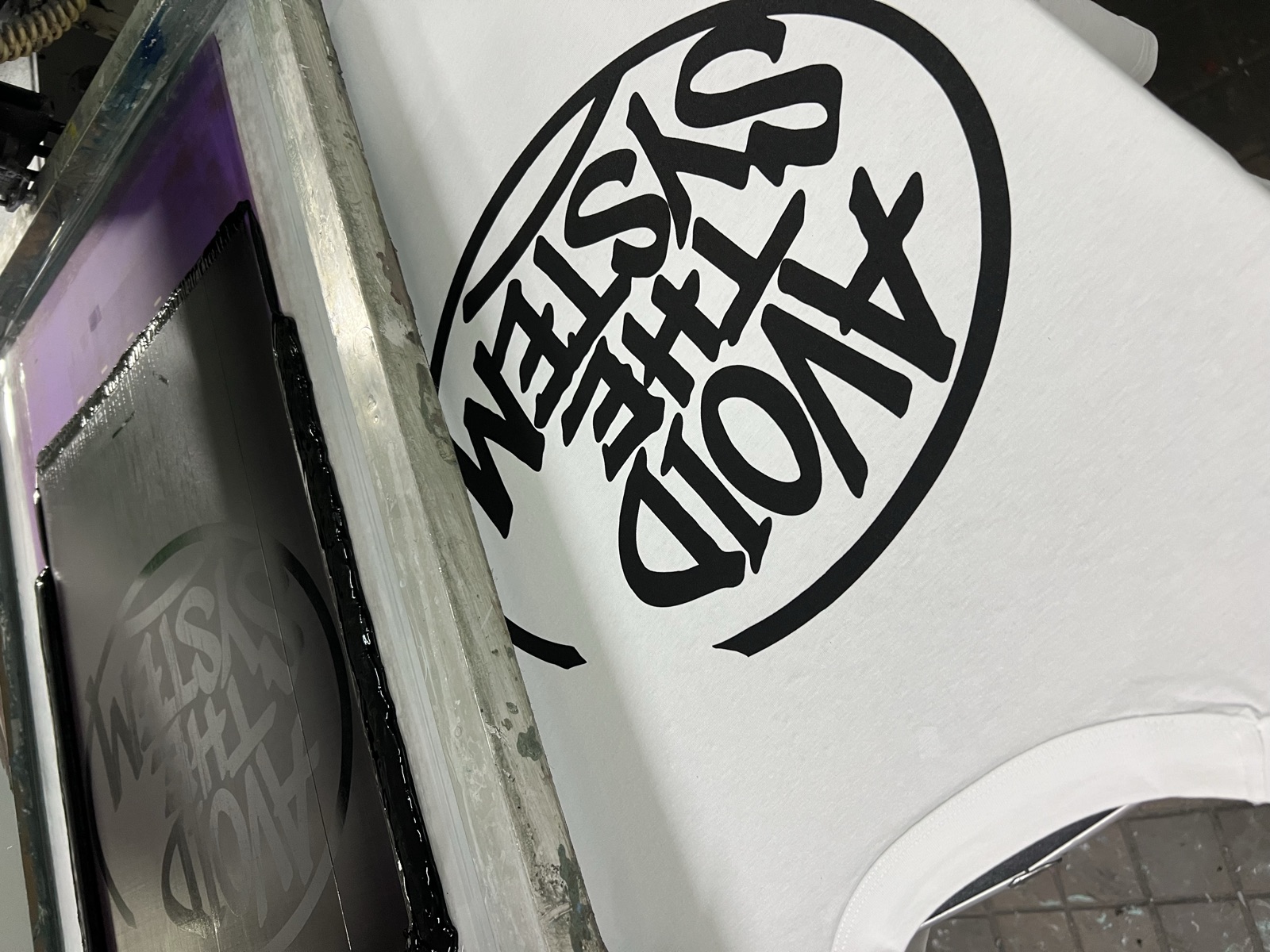 White t-shirt on screen printing press with bold black Avoid The System circular logo being applied